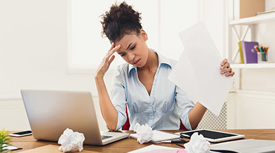 A visibly stressed woman sits in front of a laptop surrounded by untidy documents and screwed up paper.